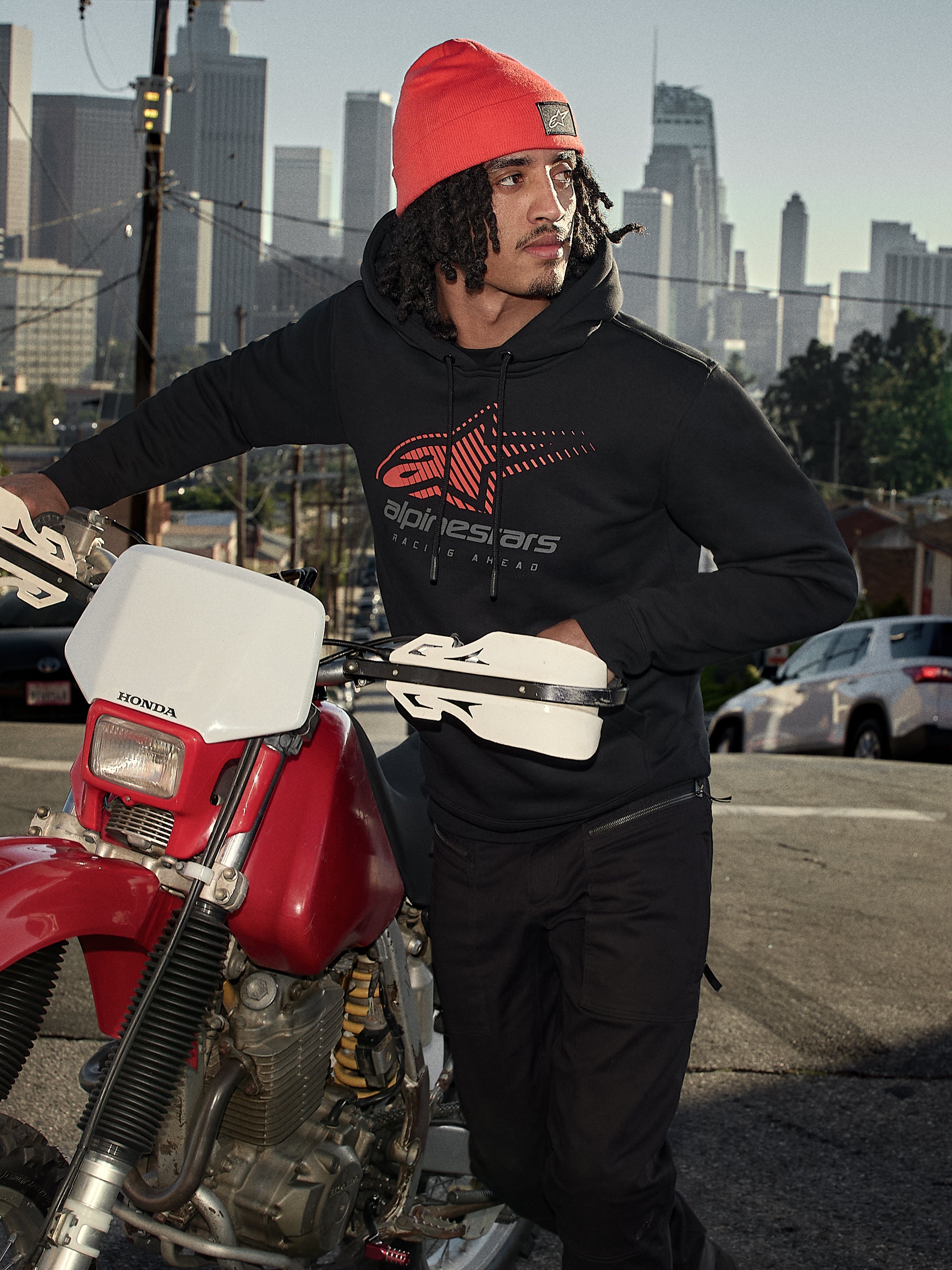 Sweat à capuche Alpinestars Pulse, noir, polaire à capuche avec logo Astars Combo rouge et gris, porté par un homme avec un bonnet orange debout à côté d'une moto Honda rouge dans un cadre urbain, streetwear décontracté inspiré des sports mécaniques.
