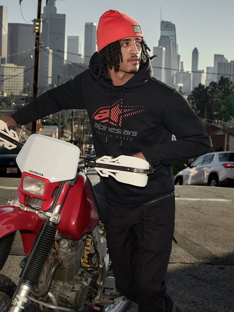 Alpinestars Pulse Hoodie, Black, hooded pullover fleece with red and grey Astars Combo Logo, worn by a man in an orange beanie standing next to a red Honda motorcycle in a city setting, casual motorsport streetwear