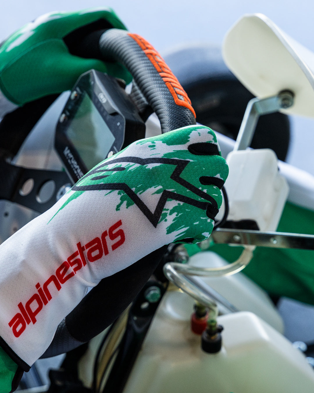 Gants Alpinestars Tech-1 K V3 en blanc, vert et rouge, mains d'un pilote serrant un volant de karting, avec un motif d'éclaboussure sublimé vert, logo étoile noir et texte Alpinestars rouge sur la manchette, équipement de karting professionnel.