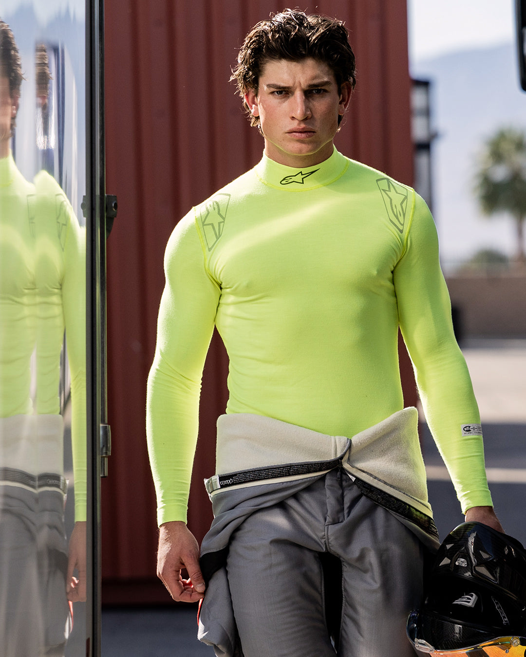 Haut à manches longues Alpinestars ZX Evo V3, sous-vêtement technique pour la course automobile, jaune fluo, porté par un pilote avec une combinaison de course grise nouée autour de la taille, tenant un casque noir, arborant le logo Alpinestars sur le col et les épaules, équipement technique anti-humidité pour le sport automobile