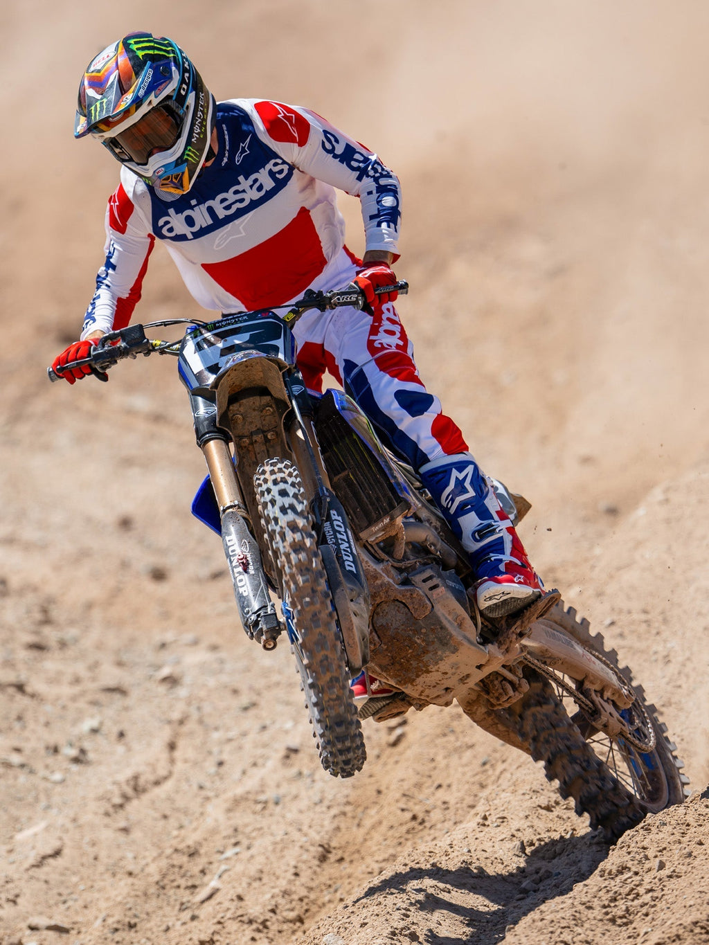 Pilote de motocross en action sur une piste de terre, portant le maillot Limited Edition Techstar Fortitude en rouge, blanc et bleu, coordonné avec un pantalon et des bottes assortis, avec logo Alpinestars, tenue de course tout-terrain professionnelle