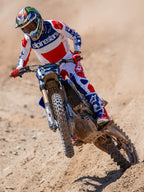 Pilote de motocross en action sur une piste de terre, portant le maillot Limited Edition Techstar Fortitude en rouge, blanc et bleu, coordonné avec un pantalon et des bottes assortis, avec logo Alpinestars, tenue de course tout-terrain professionnelle