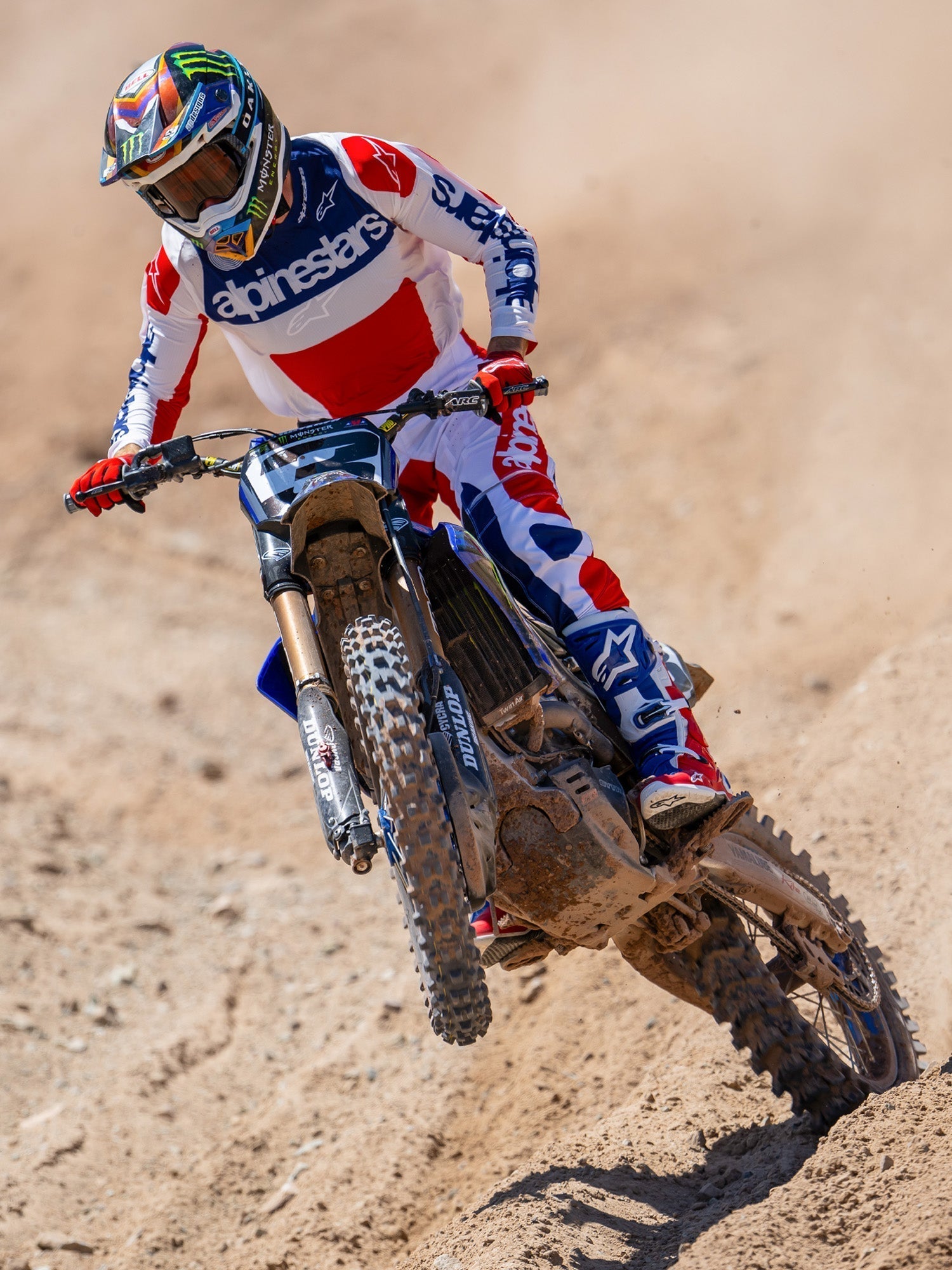 Pantalon Alpinestars Techstar Fortitude Édition Limitée, pantalon de motocross, rouge vif, blanc et bleu, porté par un pilote en action sur un terrain de terre, avec maillot et bottes assortis, équipement de course tout-terrain haute performance.
