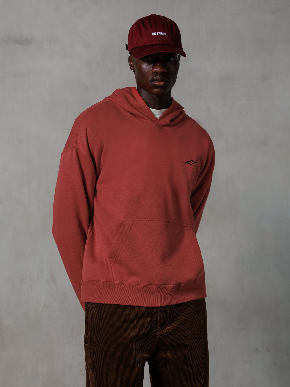 Sweat à capuche Alpinestars Essentials Ageless, streetwear décontracté, rouge brique, petit logo noir sur la poitrine, poche kangourou à l'avant, porté avec une casquette ASTARS rouge assortie et un pantalon en velours côtelé marron, design épuré et minimaliste
