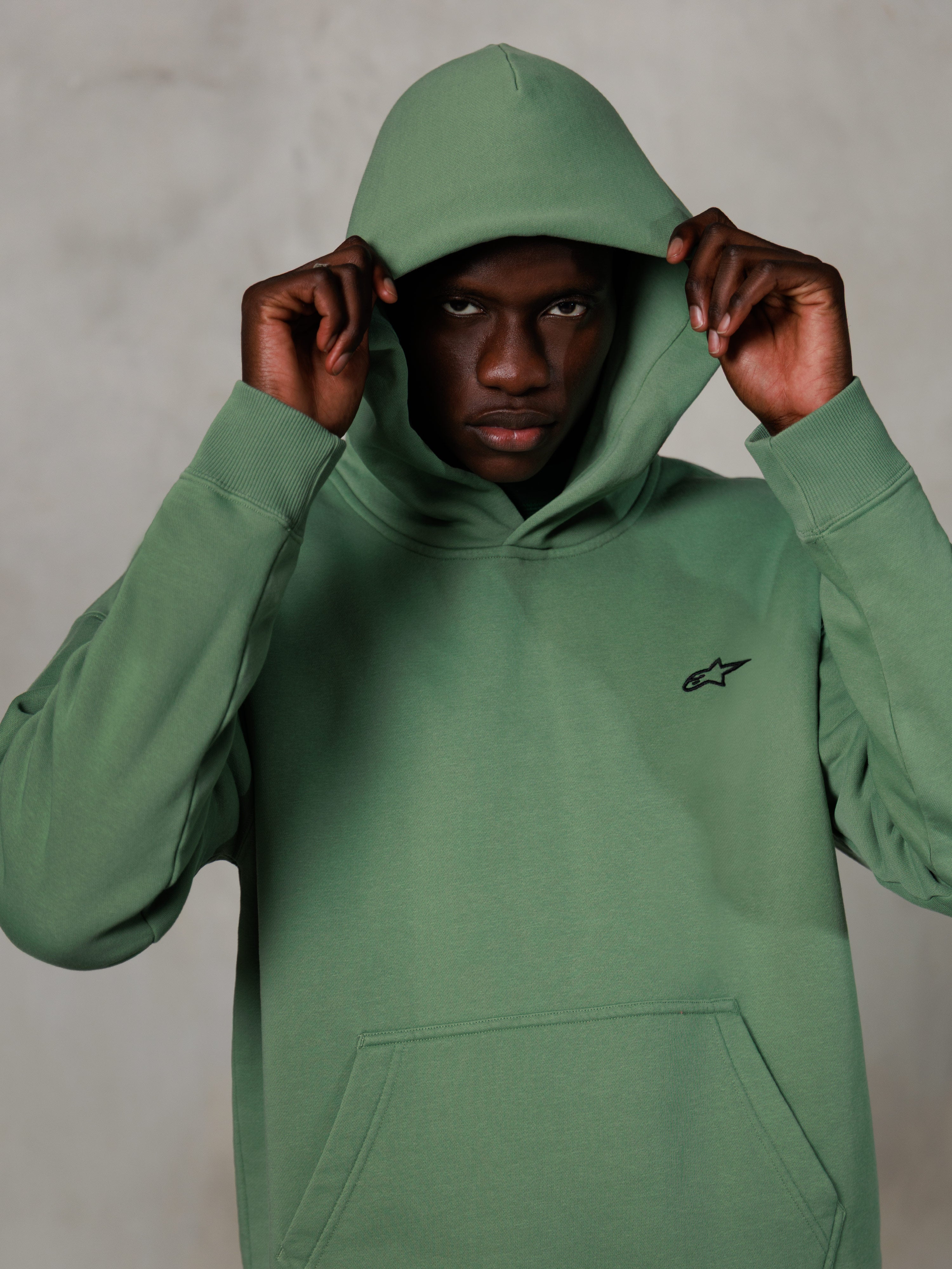 Sweat à capuche Alpinestars Essentials Ageless, streetwear décontracté, vert cactus avec petit logo noir sur la poitrine, mannequin portant le sweat avec la capuche relevée, poche kangourou frontale, design décontracté inspiré du sport automobile