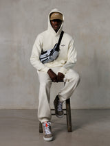 Sweat à capuche Alpinestars Essentials Ageless, Off-White, porté par un mannequin assis sur un tabouret avec un pantalon de jogging en molleton assorti, une casquette ASTARS beige et un sac bandoulière, streetwear décontracté inspiré des sports mécaniques