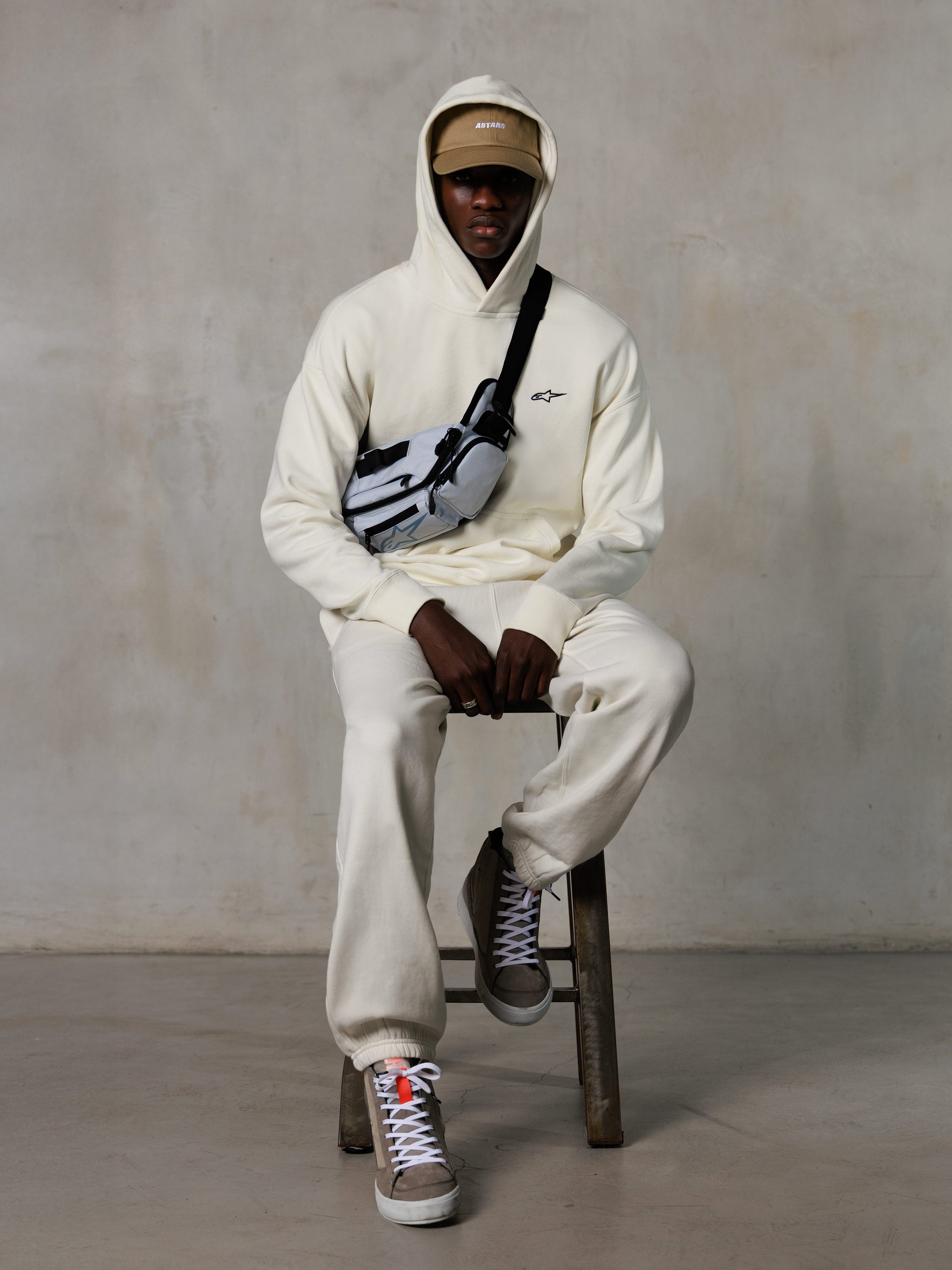 Sweat à capuche Alpinestars Essentials Ageless, Off-White, porté par un mannequin assis sur un tabouret avec un pantalon de jogging en molleton assorti, une casquette ASTARS beige et un sac bandoulière, streetwear décontracté inspiré des sports mécaniques