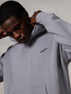 Sweat à capuche Alpinestars Essentials Ageless, Silver, gros plan d'un homme ajustant la capuche du sweat-shirt gris, petit logo Alpinestars noir sur la poitrine, vêtement décontracté haut de gamme inspiré du sport automobile