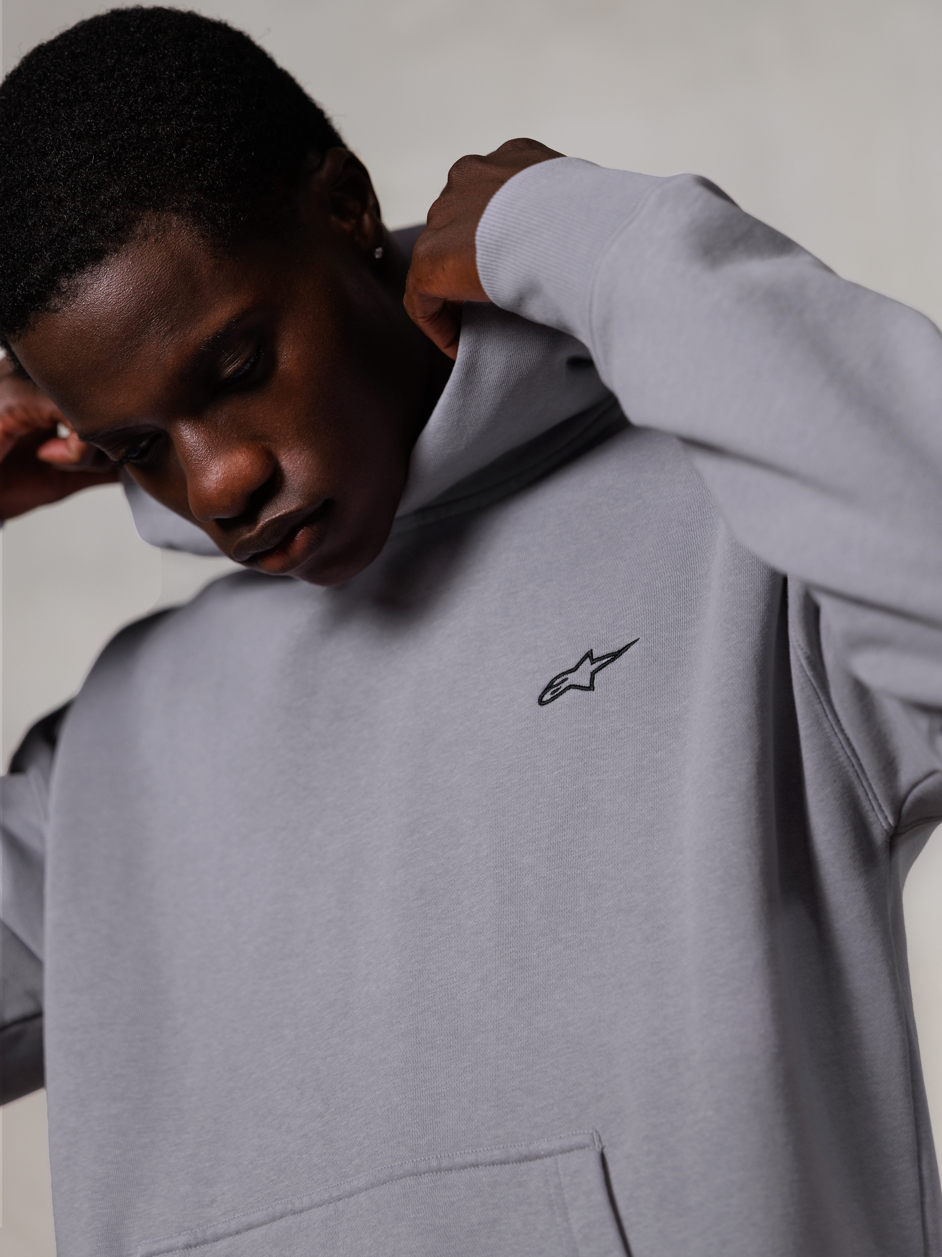Sweat à capuche Alpinestars Essentials Ageless, Silver, gros plan d'un homme ajustant la capuche du sweat-shirt gris, petit logo Alpinestars noir sur la poitrine, vêtement décontracté haut de gamme inspiré du sport automobile