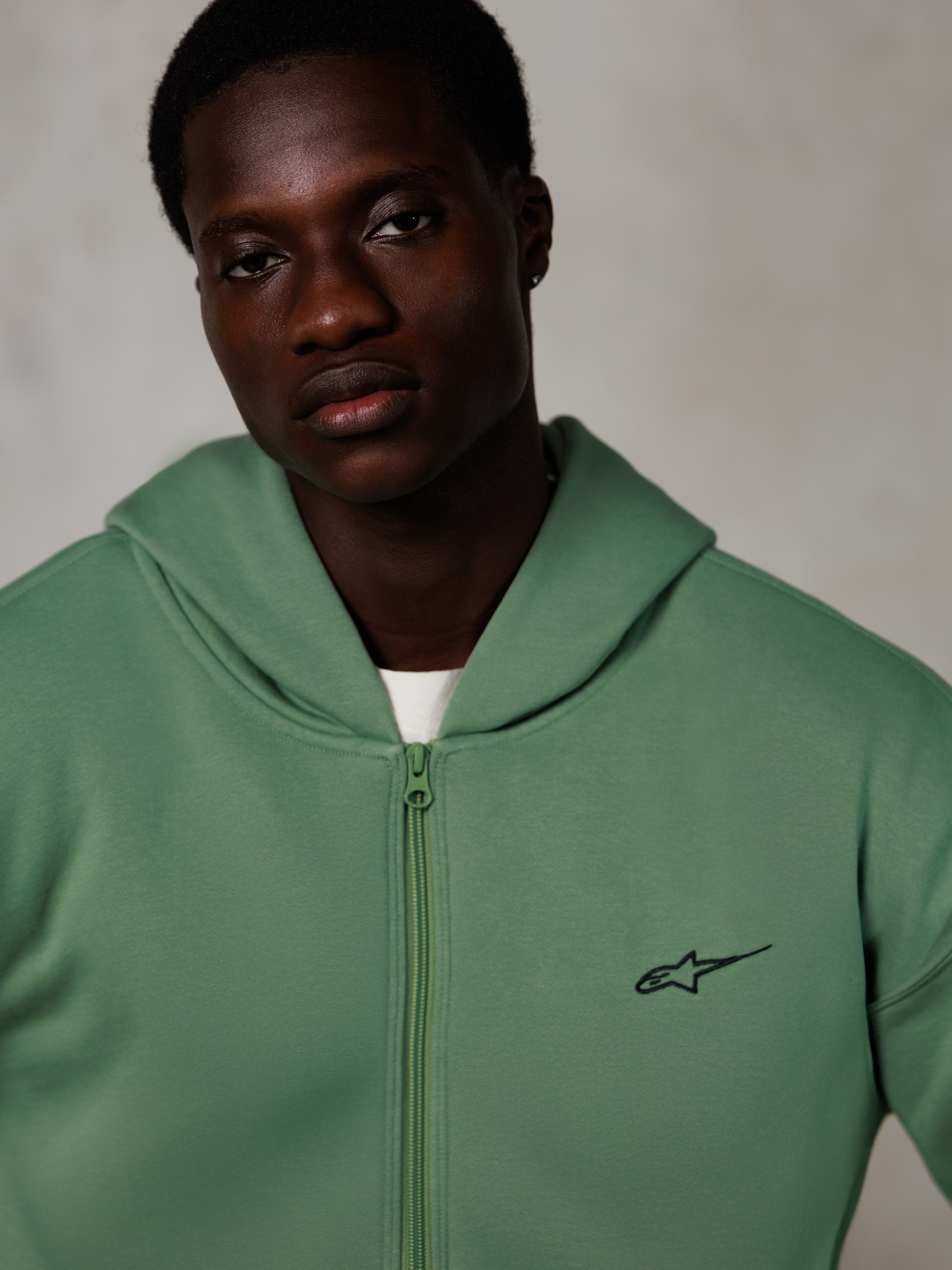 Sweat à capuche zippé Alpinestars Essentials Ageless, portrait lifestyle, vert cactus avec petit logo noir, fermeture éclair sur le devant, texture en polaire douce, streetwear décontracté inspiré du sport automobile