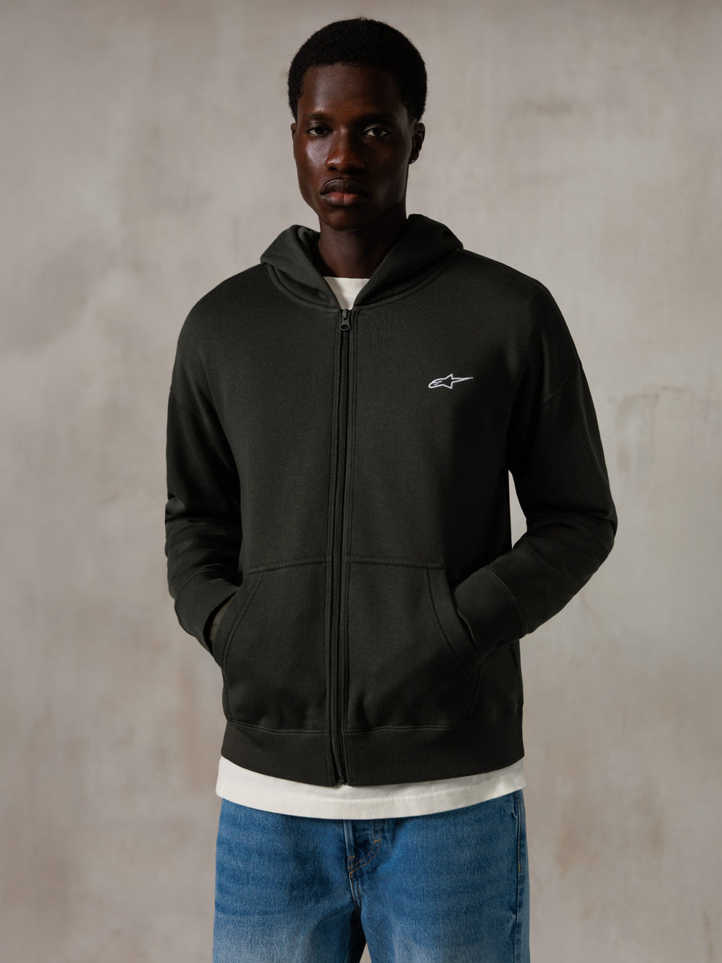 Sweat à capuche zippé Alpinestars Essentials Ageless, Off Black, sweat à capuche entièrement zippé avec un petit logo Alpinestars blanc sur la poitrine, coupe décontractée, porté par un mannequin avec un jean bleu et un t-shirt blanc, style streetwear décontracté inspiré du sport automobile