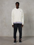 Sweat à capuche zippé Alpinestars Essentials Ageless, style streetwear décontracté, blanc cassé avec logo noir discret sur la poitrine, fermeture éclair intégrale, design à capuche, coupe décontractée, porté avec un pantalon de jogging noir et des baskets