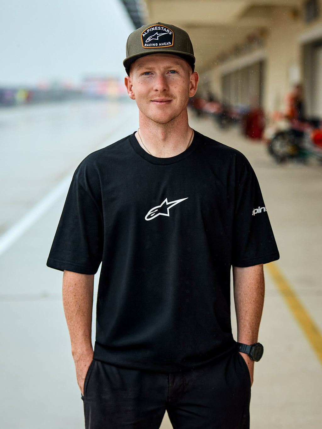 Homme portant le t-shirt Alpinestars Frontal CSF, t-shirt à manches courtes noir, logo central Astars blanc, casquette Alpinestars vert olive, streetwear décontracté d'inspiration motorsport