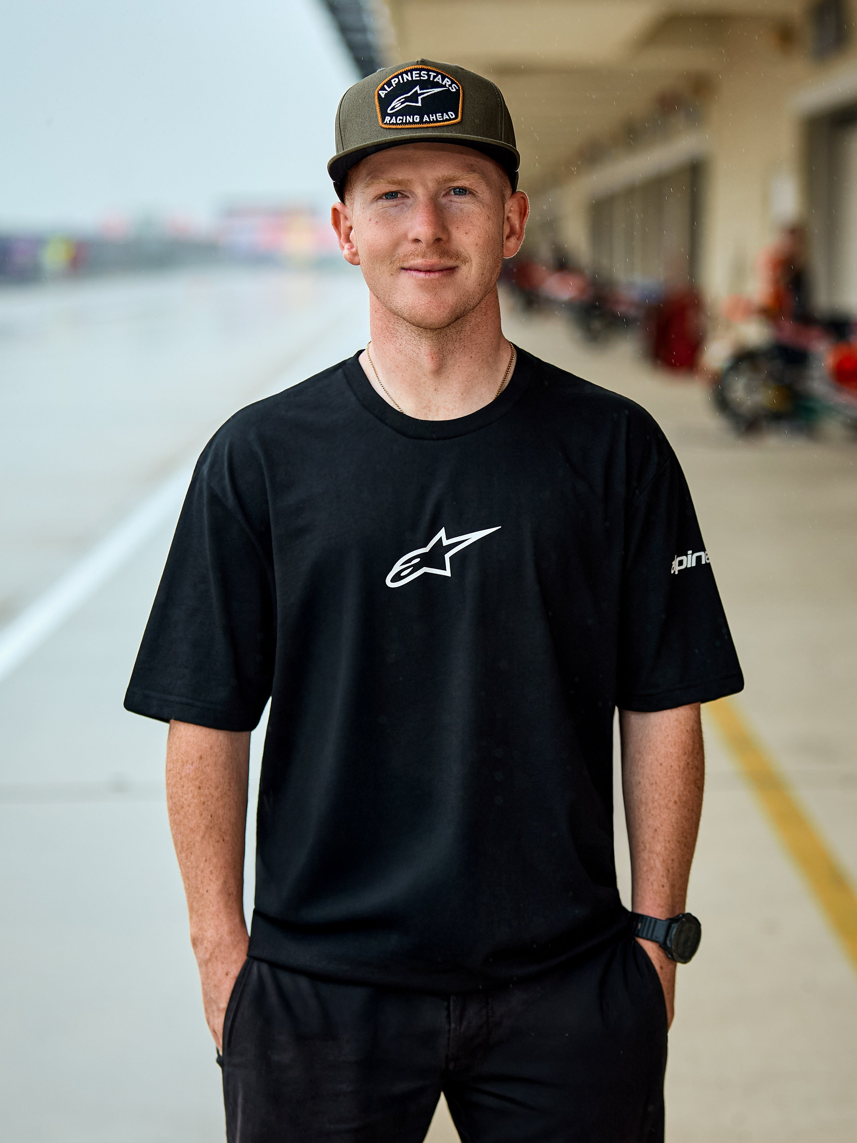 Homme portant le t-shirt Alpinestars Frontal CSF, t-shirt à manches courtes noir, logo central Astars blanc, casquette Alpinestars vert olive, streetwear décontracté d'inspiration motorsport