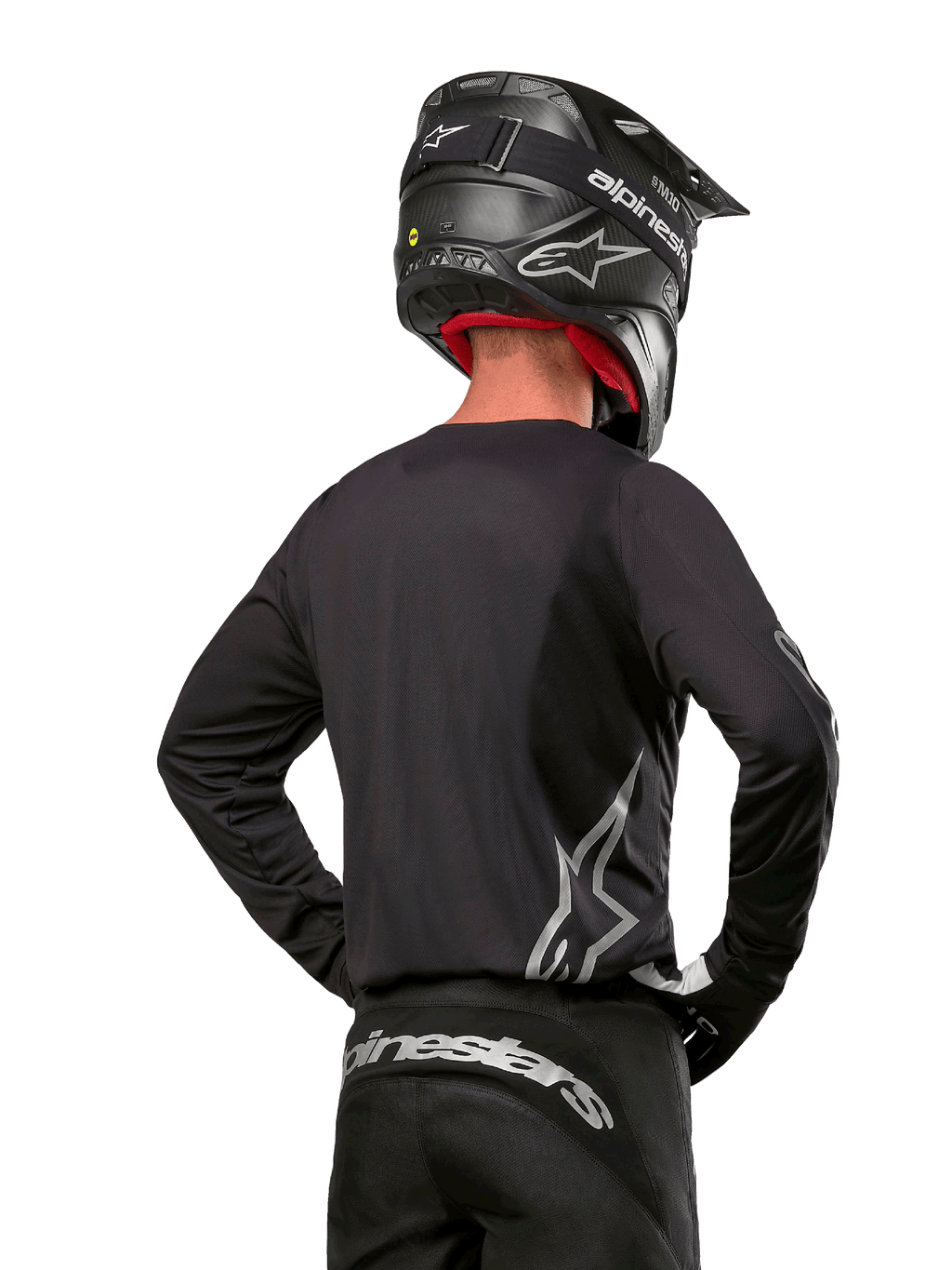 Pilote de motocross vu de dos portant le maillot Alpinestars 2024 Fluid Graphite en Noir Argent, avec un grand logo argenté sur le côté, associé à un casque noir en fibre de carbone et un pantalon noir, équipement de course tout-terrain professionnel