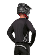 Pilote de motocross vu de dos portant le maillot Alpinestars 2024 Fluid Graphite en Noir Argent, avec un grand logo argenté sur le côté, associé à un casque noir en fibre de carbone et un pantalon noir, équipement de course tout-terrain professionnel