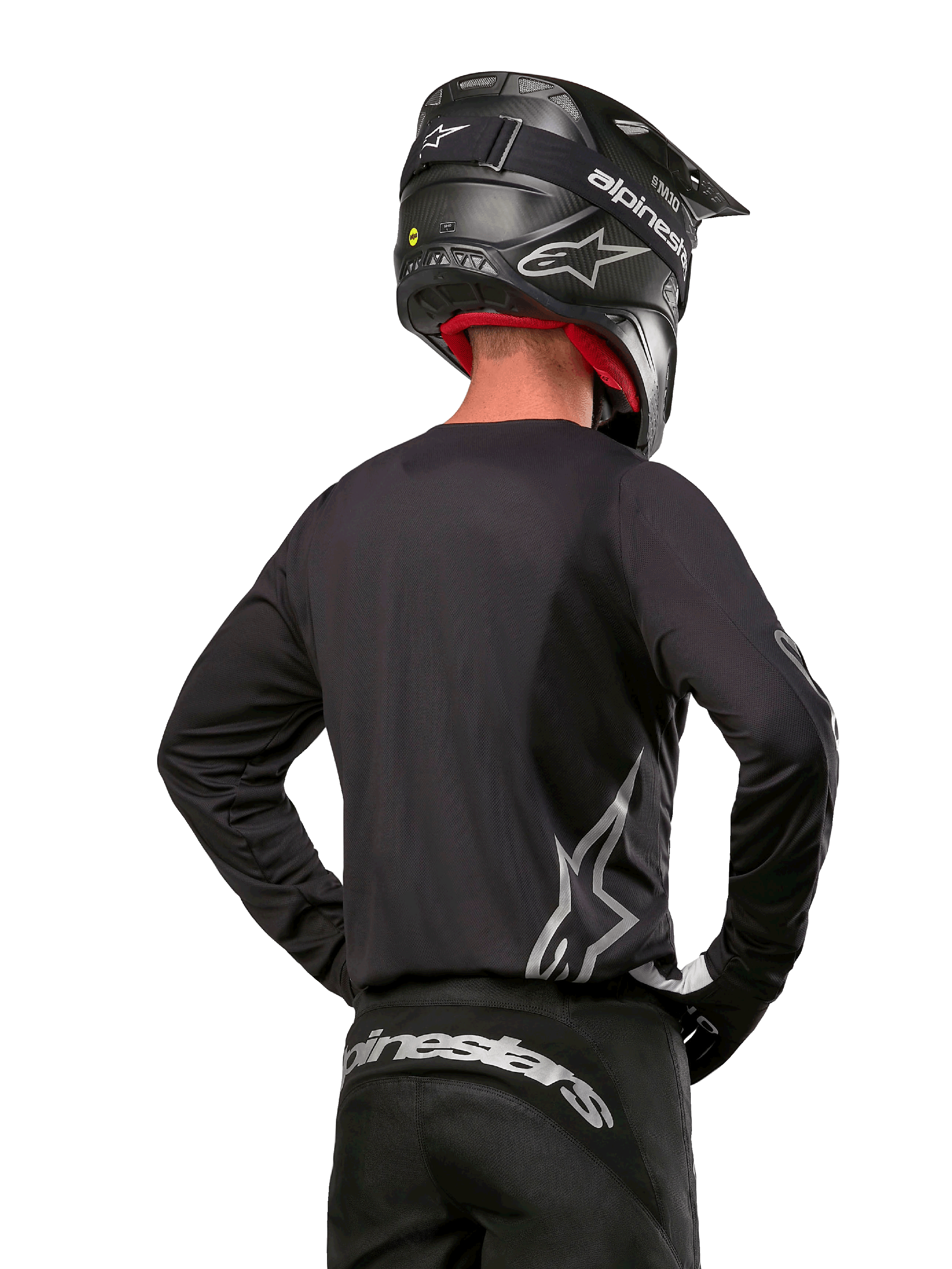 Pilote de motocross vu de dos portant le maillot Alpinestars 2024 Fluid Graphite en Noir Argent, avec un grand logo argenté sur le côté, associé à un casque noir en fibre de carbone et un pantalon noir, équipement de course tout-terrain professionnel