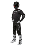 Alpinestars 2024 Fluid Graphite Gear Set, tenue de motocross complète, noir avec logos gris et blancs, comprenant un maillot et un pantalon portés avec un casque noir assorti, des gants et des bottes blancs et noirs, conçu pour la protection tout-terrain