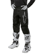 Pantalon Alpinestars Fluid Graphite 2024, pantalon de motocross, gris et noir, porté avec des bottes et des gants de motocross blancs et noirs, arborant un grand logo Alpinestars argenté sur la cuisse, coupe sportive athlétique pour la conduite tout-terrain