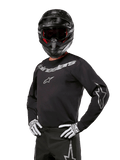 Alpinestars 2024 Fluid Graphite Jersey, maillot de motocross noir et argent, coupe performance à manches longues, porté avec un casque noir et des gants blancs, avec logos Alpinestars argentés, équipement de course tout-terrain