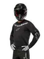 Alpinestars 2024 Fluid Graphite Jersey, maillot de motocross noir et argent, coupe performance à manches longues, porté avec un casque noir et des gants blancs, avec logos Alpinestars argentés, équipement de course tout-terrain