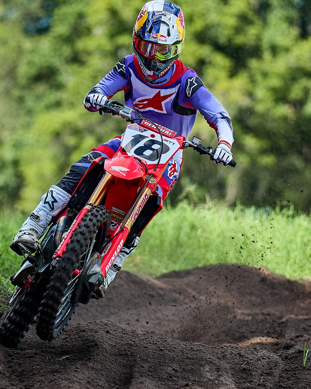 Pilote de motocross sur une moto Honda rouge, portant le maillot Alpinestars Fluid Wurx 2025 violet et rouge vif, pantalon assorti, bottes blanches et casque Red Bull, négociant un virage sur une piste de terre