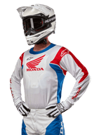 Pilote de motocross portant le maillot Honda Racer Iconic en blanc, bleu vif et rouge vif, avec un grand logo d'aile Honda sur la poitrine, associé à un casque Alpinestars blanc et des gants blancs et noirs assortis, équipement coordonné pour la performance tout-terrain