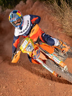 Pilote de motocross penché dans un virage sur une moto orange, portant le maillot et le pantalon Alpinestars Pro-Dura en Bleu Navy, Orange Vif et Blanc, tenue de course tout-terrain coordonnée, casque Red Bull, bottes blanches, photo d'action sur une piste de terre poussiéreuse.