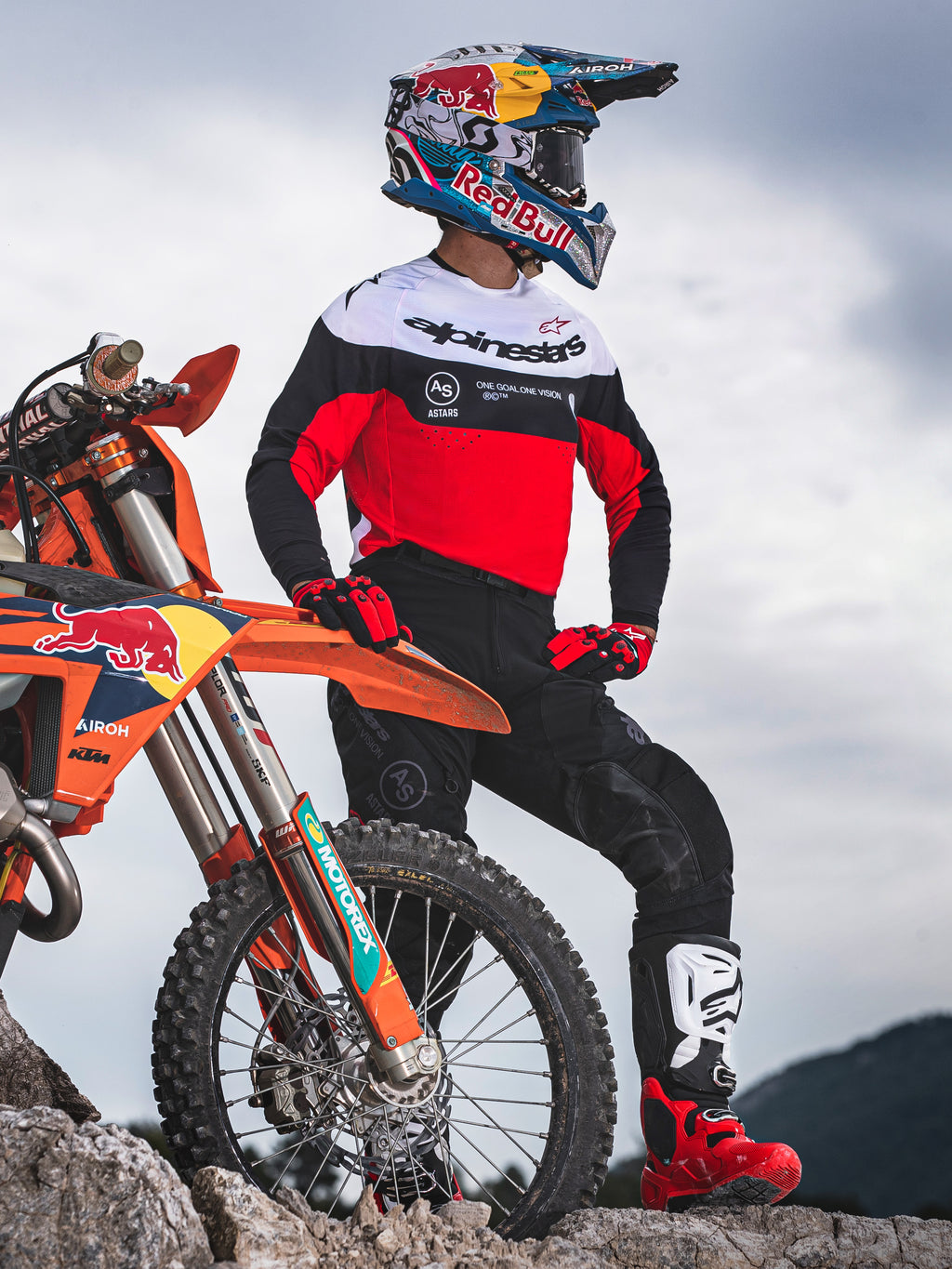 Pilote de motocross portant le maillot Alpinestars Pro-Dura noir, rouge vif et blanc, debout à côté d'une moto KTM sur un terrain rocheux, associé à un pantalon noir et des bottes de motocross rouges, cadre enduro extérieur.