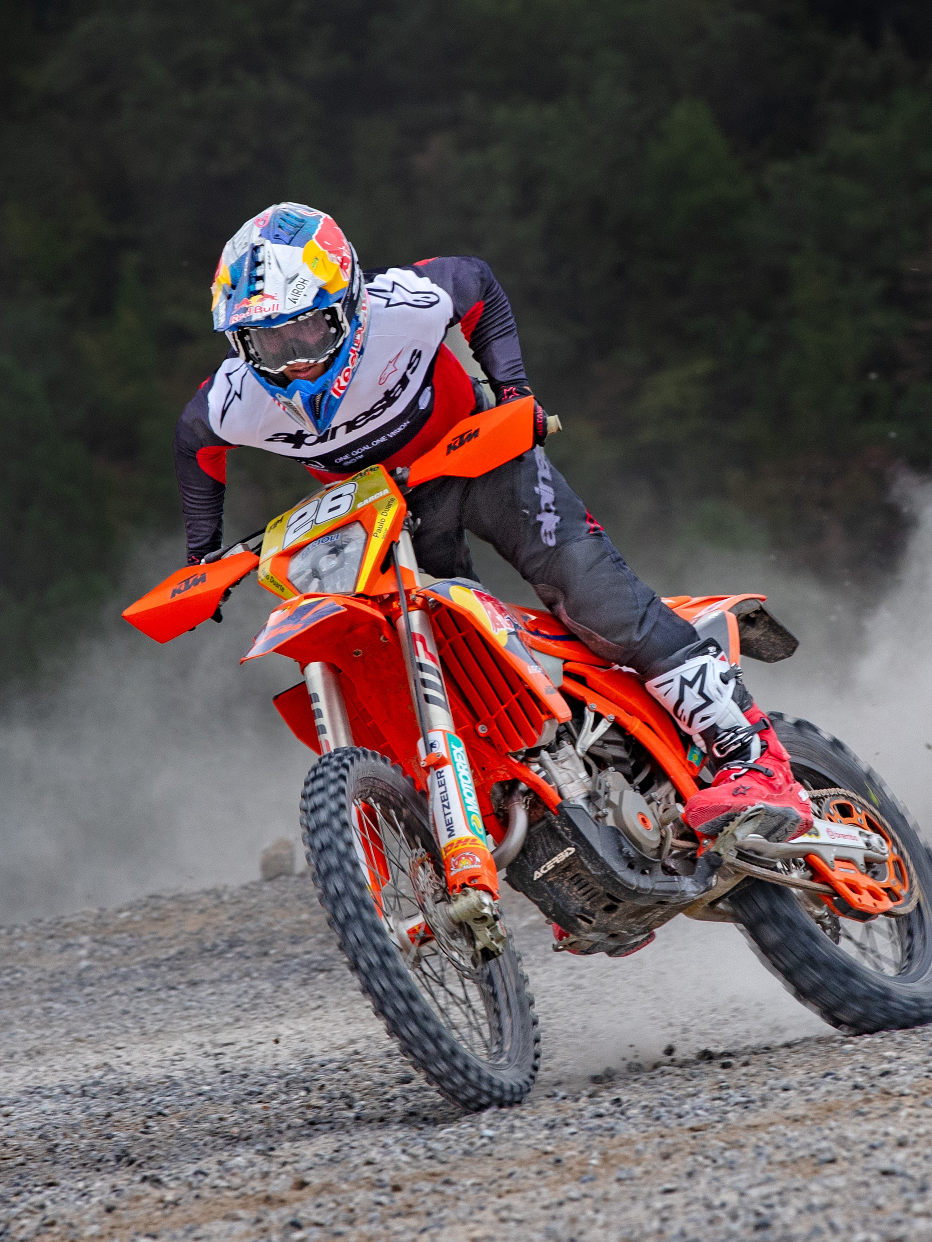 Pilote de motocross en action portant un maillot Alpinestars Pro-Dura noir, rouge vif et blanc, pilotant une moto orange sur un sentier poussiéreux, avec un pantalon noir et des bottes rouges et blanches