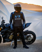 Veste Alpinestars T-Stunt Air, veste textile à coupe sportive, noir, bleu foncé et bleu vif, portée par un pilote à côté d'une moto Ducati, avec un casque en fibre de carbone et une tenue de moto noire, dans un paysage désertique au coucher du soleil.