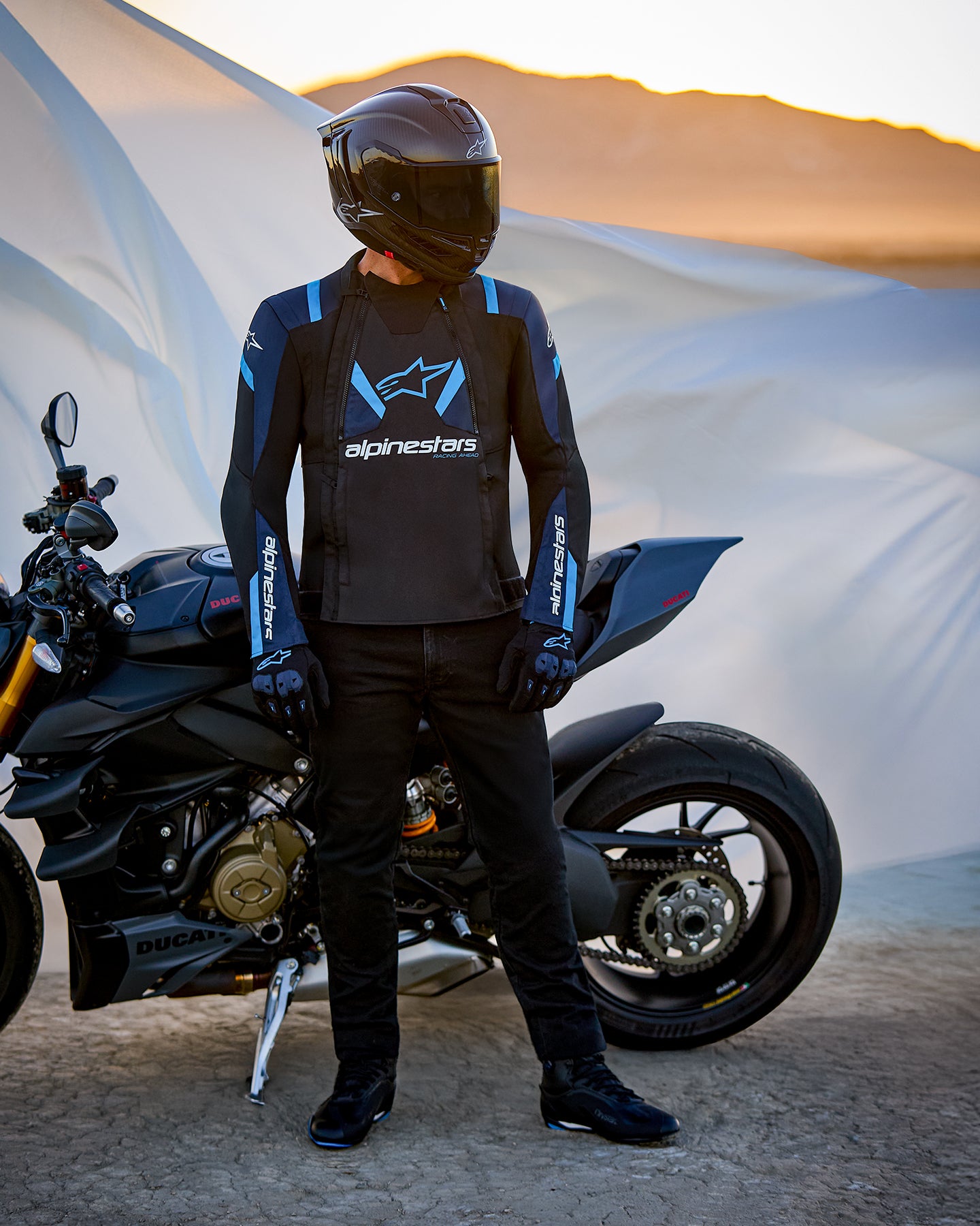 Veste Alpinestars T-Stunt Air, veste textile à coupe sportive, noir, bleu foncé et bleu vif, portée par un pilote à côté d'une moto Ducati, avec un casque en fibre de carbone et une tenue de moto noire, dans un paysage désertique au coucher du soleil.