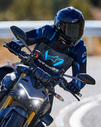 Pilote de moto s'inclinant dans un virage portant la veste Alpinestars T-Stunt Air en noir, bleu foncé et bleu vif, avec un casque et des gants noirs, photo d'action sur route goudronnée, équipement de conduite sportive haute performance