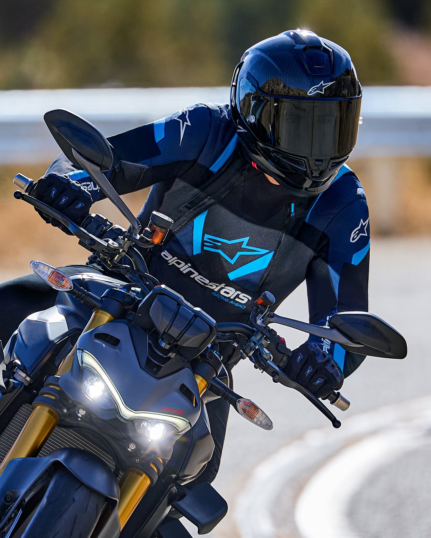 Pilote de moto s'inclinant dans un virage portant la veste Alpinestars T-Stunt Air en noir, bleu foncé et bleu vif, avec un casque et des gants noirs, photo d'action sur route goudronnée, équipement de conduite sportive haute performance