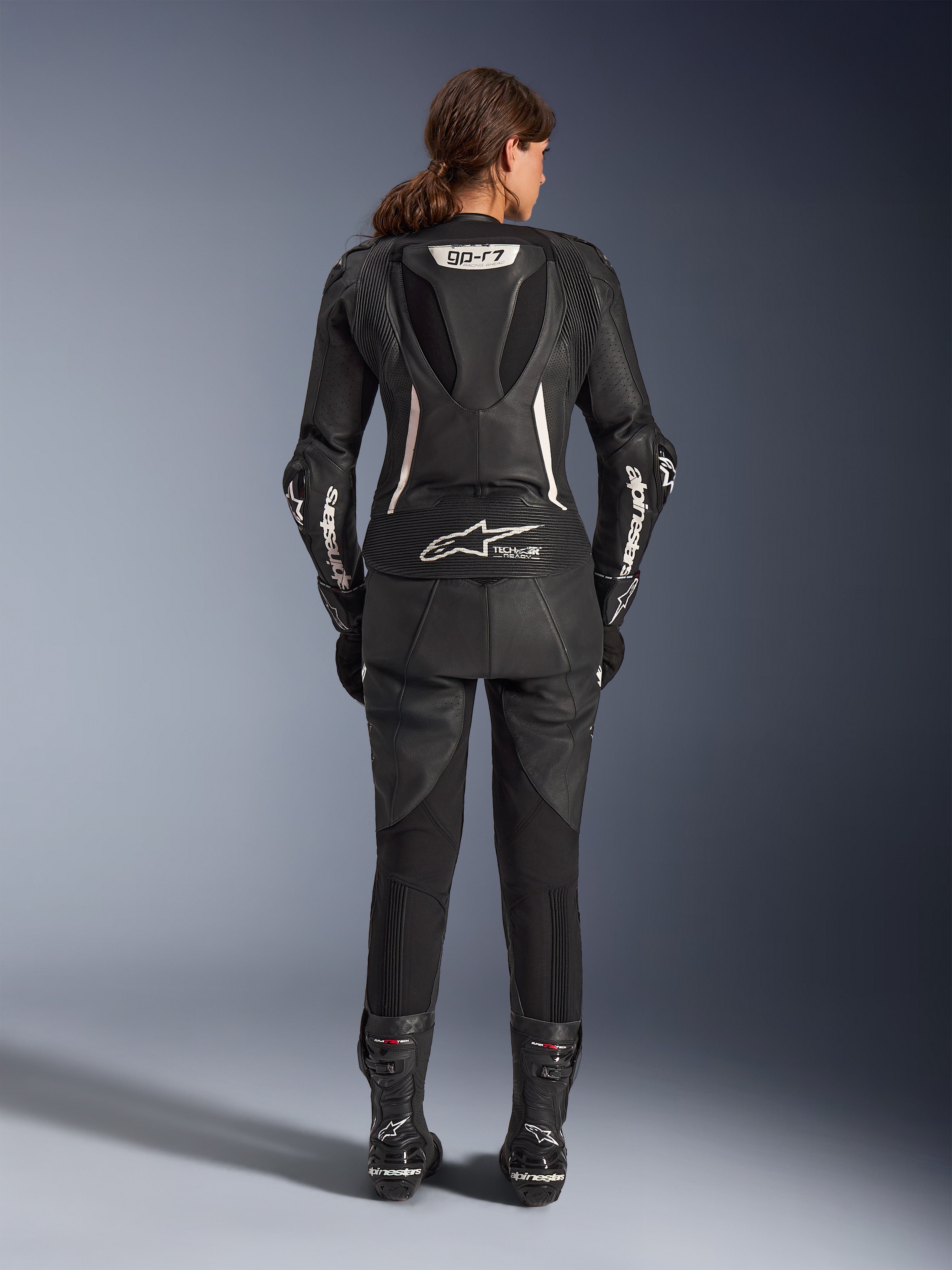 Combinaison en cuir 2 pièces Alpinestars Stella GP-R7, combinaison moto pour femme, noire avec accents blancs, vue arrière montrant la bosse aérodynamique GP-R7 et le marquage Tech-Air Ready, logos Alpinestars blancs sur les manches, panneaux extensibles en accordéon, portée avec des bottes de course noires