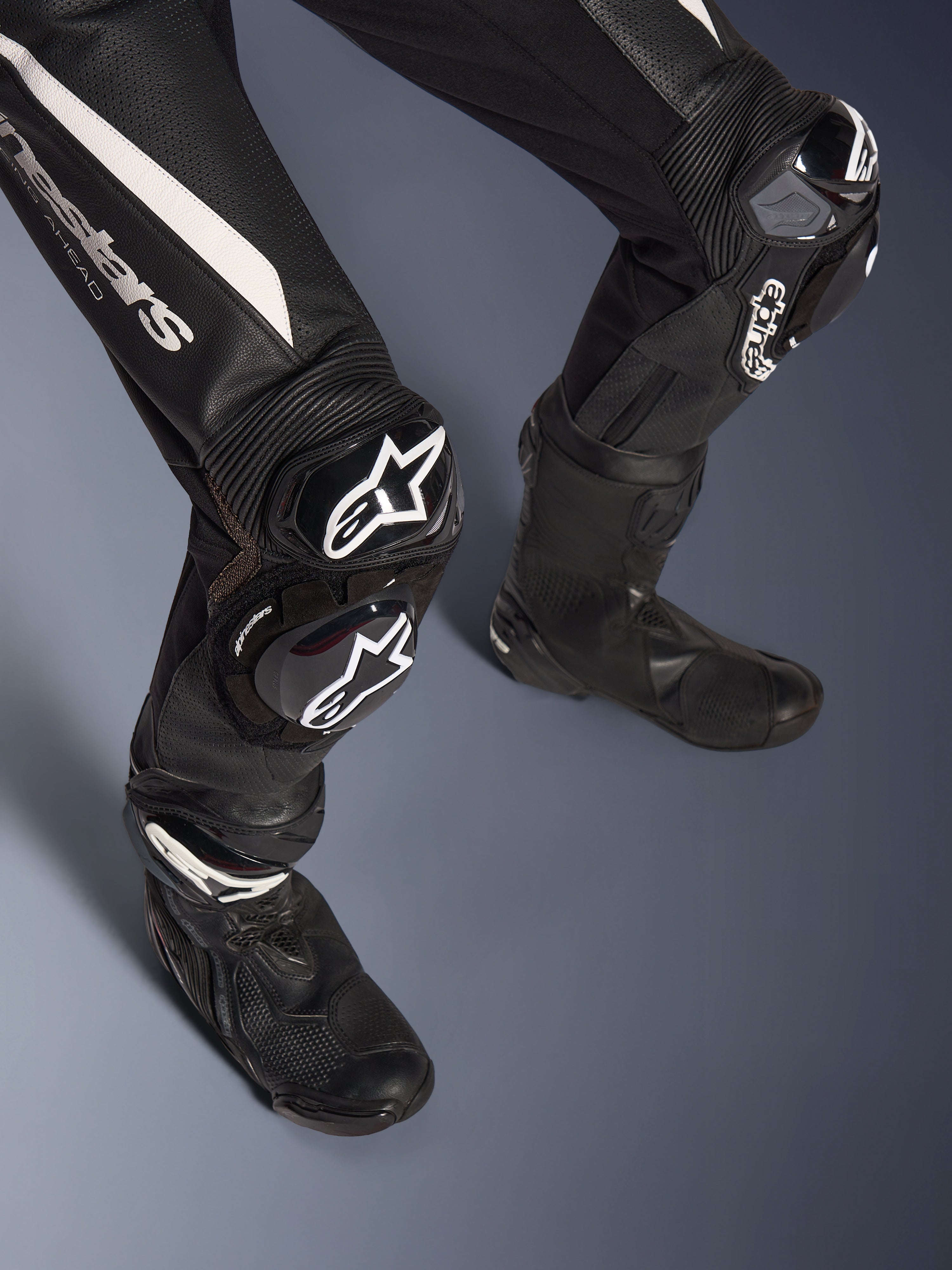 Détail des jambes de la combinaison en cuir 2 pièces Alpinestars Stella GP-R7, noir avec des accents blancs, dotée de protections de genou et de sliders GP DFS RR avec logo Alpinestars, associée à des bottes de course noires, conçue pour la performance sur piste