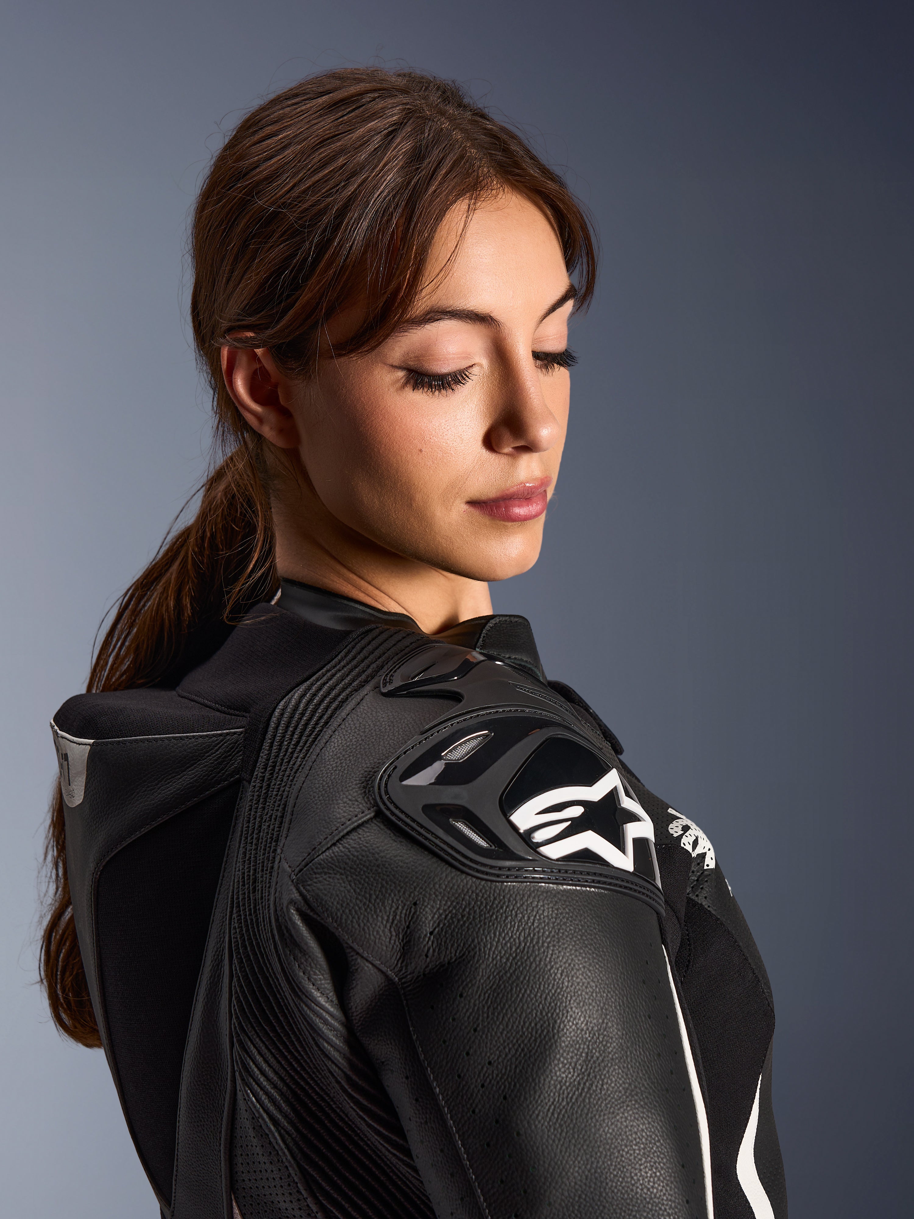 Femme portant la combinaison en cuir 2 pièces Alpinestars Stella GP-R7, vue rapprochée de l'épaule en cuir noir avec protection DFS RR et logo étoile blanc, présentant la bosse aérodynamique intégrée et les détails perforés.