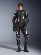 Femme portant la combinaison en cuir 2 pièces Alpinestars Stella GP-R7 en noir avec accents blancs, dotée de panneaux en cuir perforé et de logos blancs sur le torse et les genoux, assortie de gants et de bottes de course noirs, équipement professionnel de course de moto