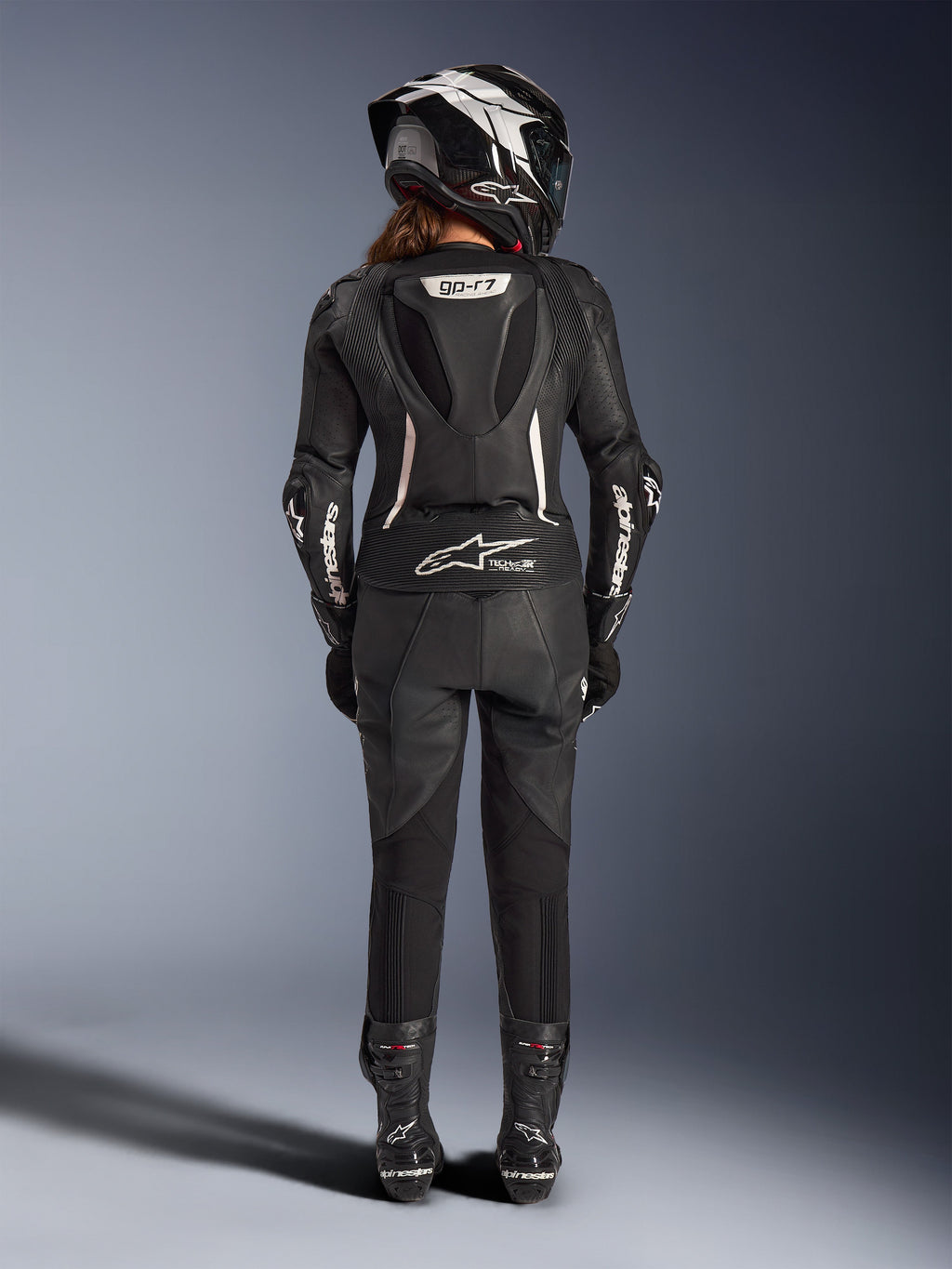 Combinaison en cuir 2 pièces Stella GP-R7 Alpinestars pour femme, noire avec accents blancs, vue arrière montrant la bosse aérodynamique Supertech R10 et le logo Tech-Air Ready, portée avec casque intégral, gants de course et bottes en cuir pour la piste et la conduite sportive