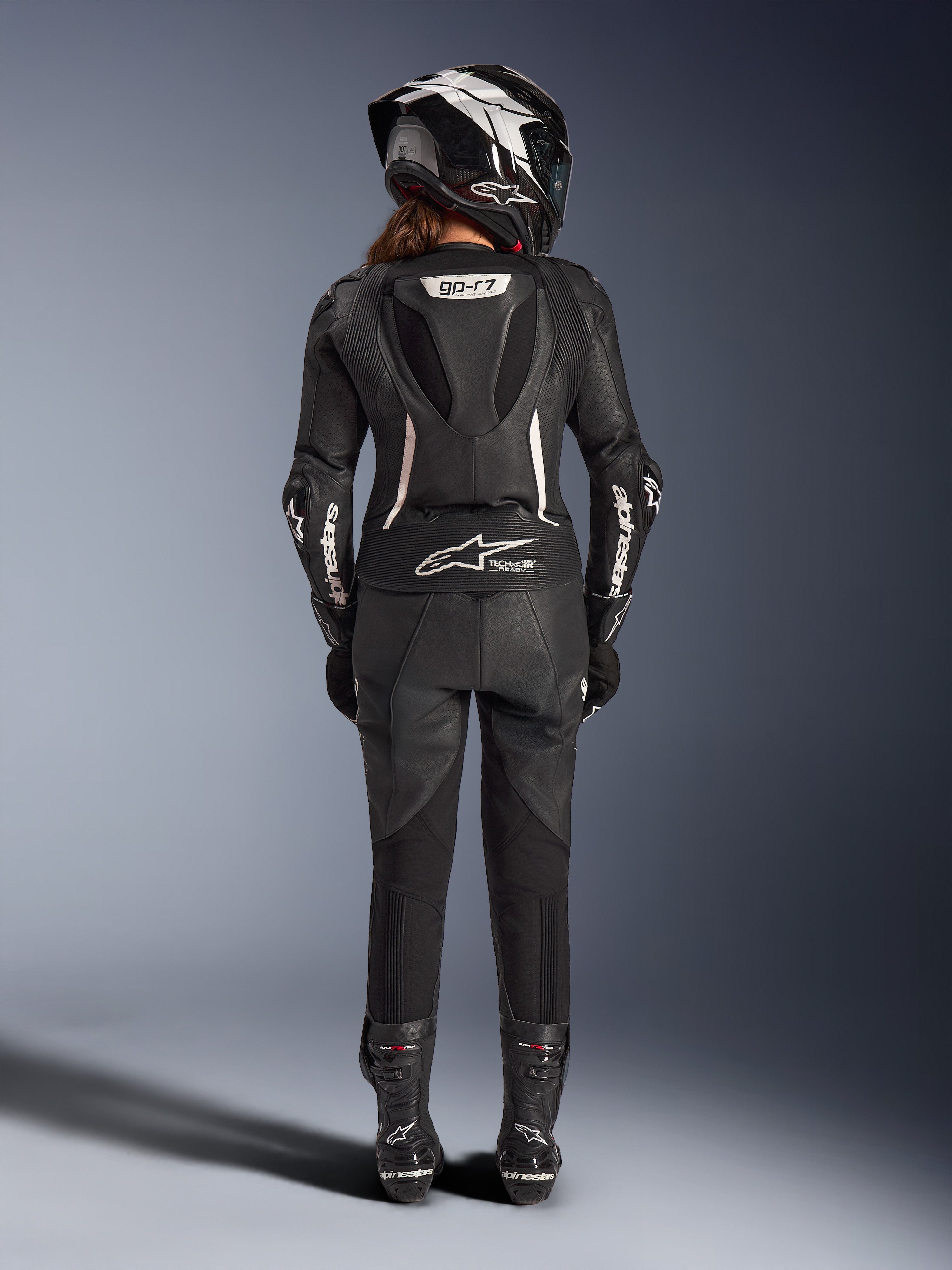 Combinaison en cuir 2 pièces Stella GP-R7 Alpinestars pour femme, noire avec accents blancs, vue arrière montrant la bosse aérodynamique Supertech R10 et le logo Tech-Air Ready, portée avec casque intégral, gants de course et bottes en cuir pour la piste et la conduite sportive