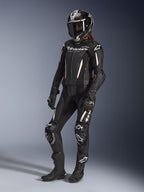 Alpinestars Stella GP-R7 2 pièces, combinaison en cuir pour femme, noire avec des accents blancs, portée par une pilote avec un casque intégral de course, des gants noirs et des bottes, présentant des panneaux en cuir perforé et le logo Alpinestars blanc, équipement de course professionnel et de performance sportive