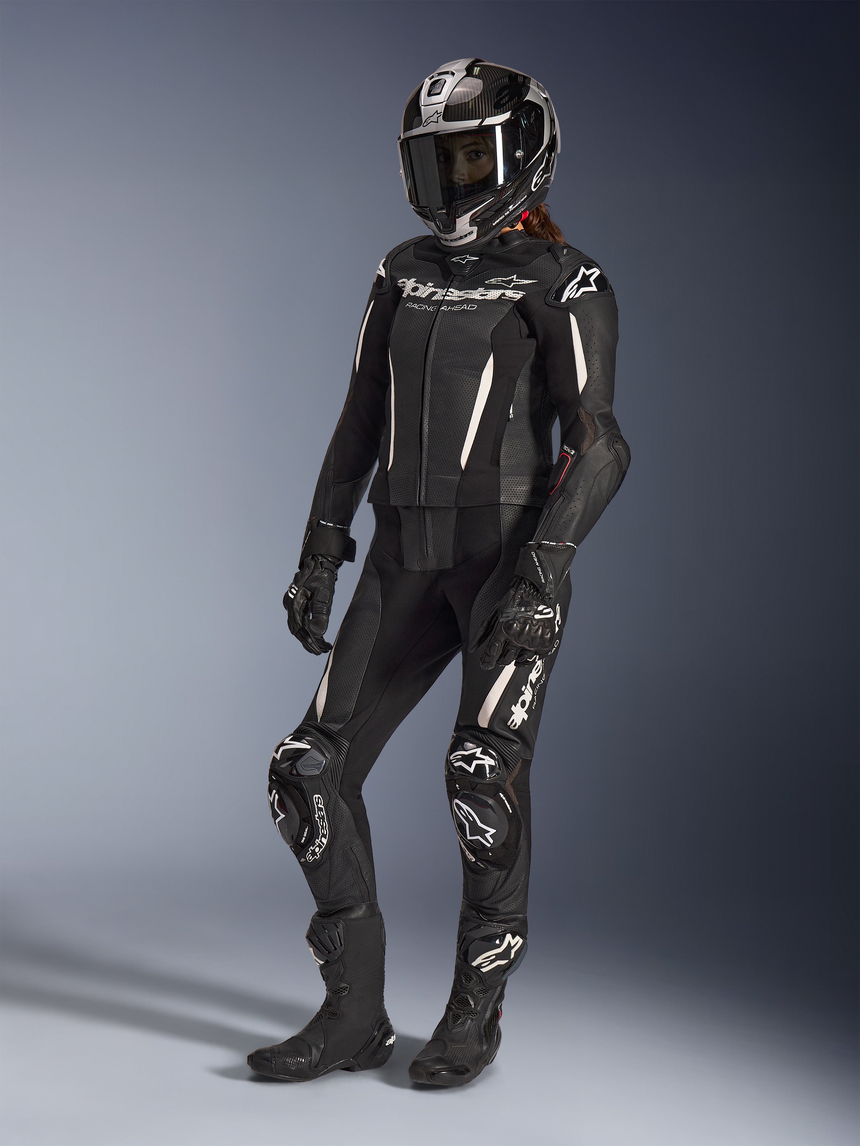 Alpinestars Stella GP-R7 2 pièces, combinaison en cuir pour femme, noire avec des accents blancs, portée par une pilote avec un casque intégral de course, des gants noirs et des bottes, présentant des panneaux en cuir perforé et le logo Alpinestars blanc, équipement de course professionnel et de performance sportive