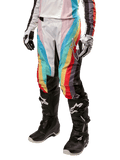 Alpinestars Stella Techstar 2024, pantalon de motocross pour femme, noir multicolore avec rayures diagonales rouges, jaunes et bleues, porté avec des bottes de moto noires et des gants blancs, avec panneaux de protection noirs aux genoux et marquage audacieux pour la course tout-terrain