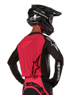 Maillot Alpinestars Supertech Ward 2024, maillot de motocross, noir et rouge berry, vue arrière montrant le panneau en maille rouge et les manches noires avec logo blanc, porté avec un casque en fibre de carbone noir et des gants, coupe de course athlétique pour la conduite tout-terrain