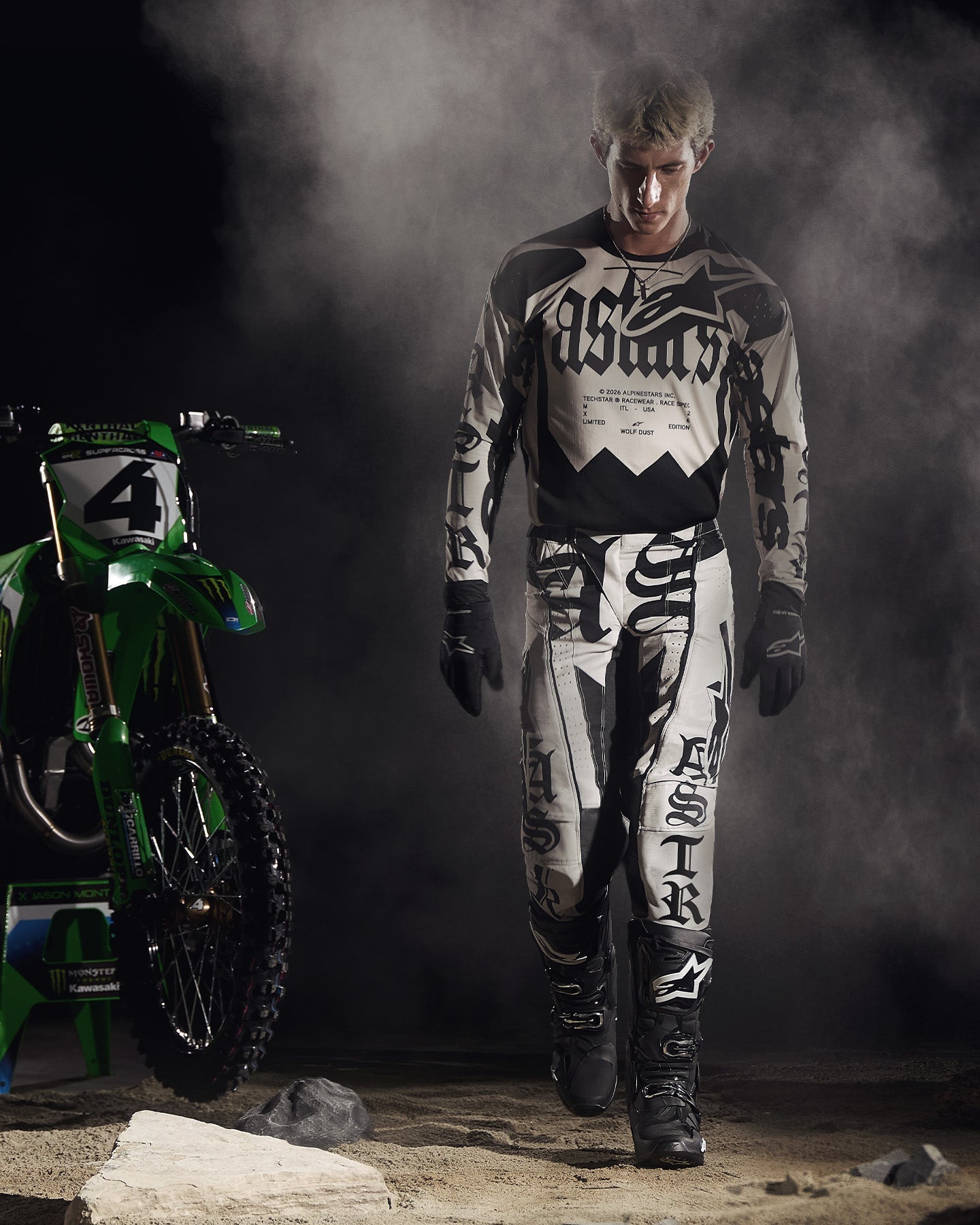 Alpinestars Limited Edition S-M7 Wolf Dust collection, Dune Black and white motocross gear on a rider next to a green bike.
