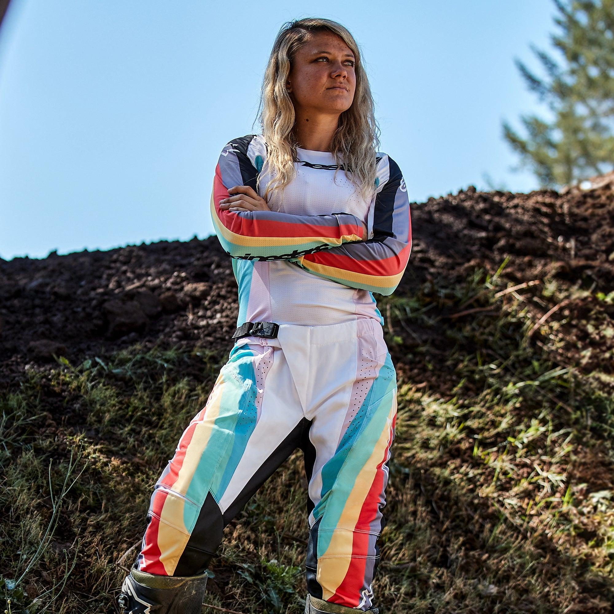 Pilote de motocross femme portant le pantalon Alpinestars Stella Techstar 2024 et le maillot assorti, design Noir Multicolore avec base blanche et rayures éclatantes bleues, jaunes et rouges, debout les bras croisés sur une piste de terre, tenue de course tout-terrain professionnelle