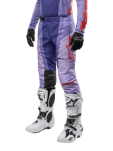 Alpinestars 2024 Techstar Ocuri Pants, pantalon de motocross, violet avec accents orange vif, logo rouge, porté avec des bottes blanches et noires et gants noirs, équipement de protection technique pour tout-terrain