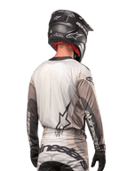 Pilote de motocross vu de dos portant le maillot Alpinestars 2024 Techstar Pneuma en Dark Sand Iron Dust Gray, associé à un casque noir à finition carbone, avec un grand logo étoile dans le dos et le marquage Alpinestars sur les manches.