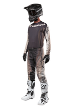 Pantalon de motocross Alpinestars Techstar Pneuma 2024, Dark Sand Iron Dust Gray, porté avec une tenue complète comprenant un maillot assorti, un casque noir, des gants noirs et des bottes blanches, avec des motifs camouflage désert ton sur ton et de grands logos.
