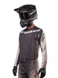 Maillot Alpinestars Techstar Pneuma 2024, maillot de motocross, sable foncé et gris fer, haut de performance à manches longues avec perforations au laser et logos blancs, porté avec un pantalon assorti et un casque noir, coupe athlétique pour le pilotage tout-terrain